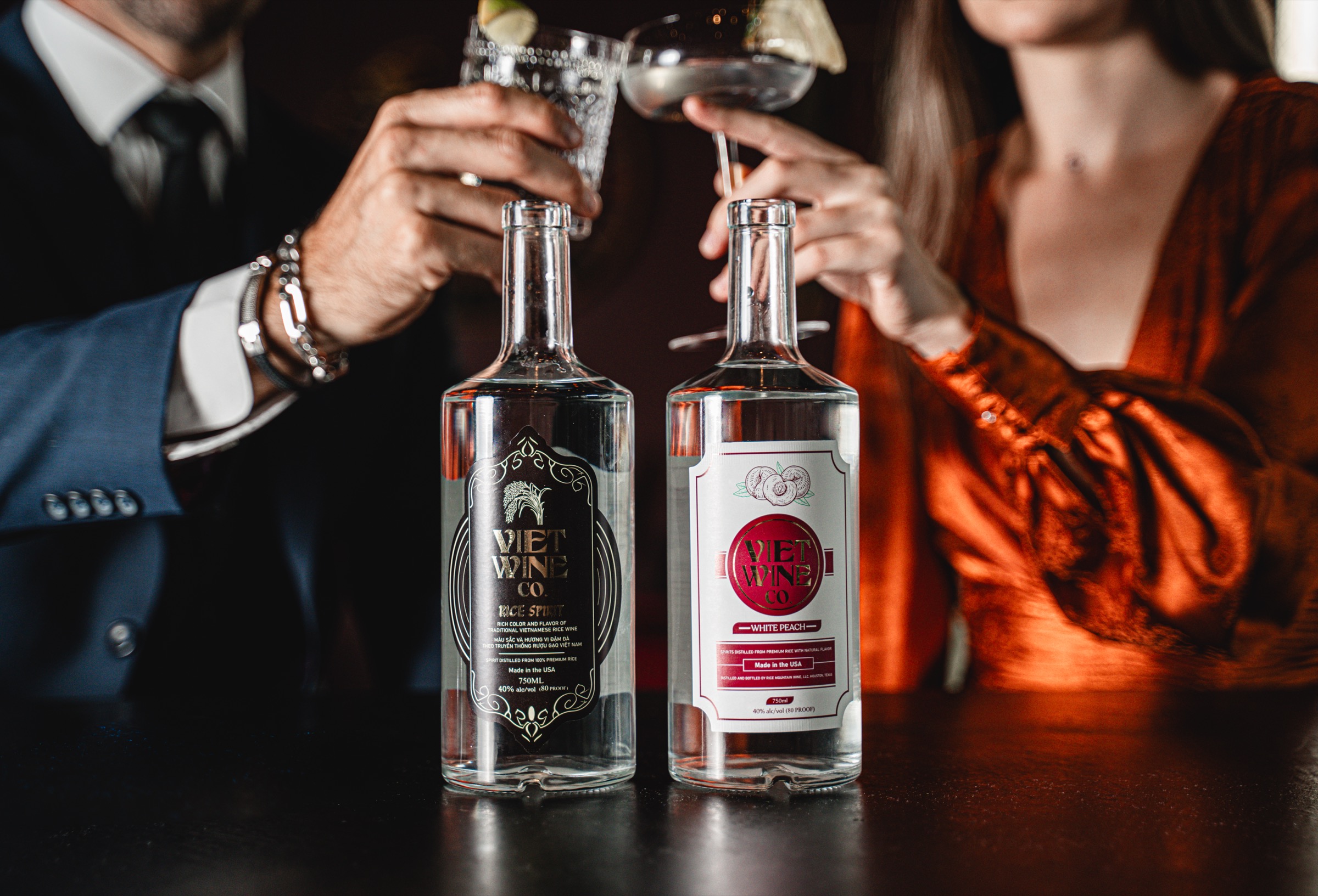 A couple toasting over bottles of Viet Wine Co. Rice Spirit and White Peach at a bar